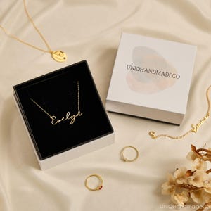May include: A gold necklace with the name Evelyn in script font inside a black and white gift box labeled UNIQHANDMADECO. Additional items include a gold necklace with a butterfly pendant, a gold chain with the name Emma, and two gold rings.