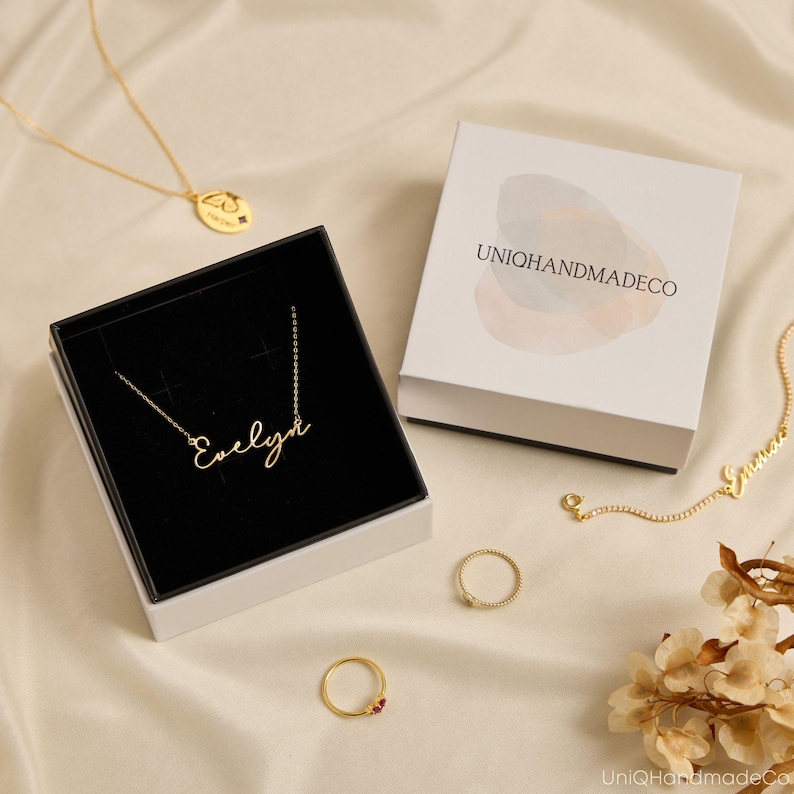 May include: A gold necklace with the name Evelyn in script font inside a black and white gift box labeled UNIQHANDMADECO. Additional items include a gold necklace with a butterfly pendant, a gold chain with the name Emma, and two gold rings.