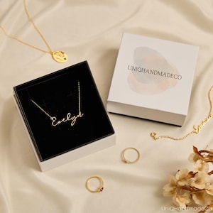 May include: A gold-colored necklace with the name "Evelyn" in cursive script is displayed in a black box. Other jewelry includes a ring with a red stone, a ring with small stones, and a necklace with a circular pendant. A white box with the logo "UNIOQHANDMADECO" is also present.