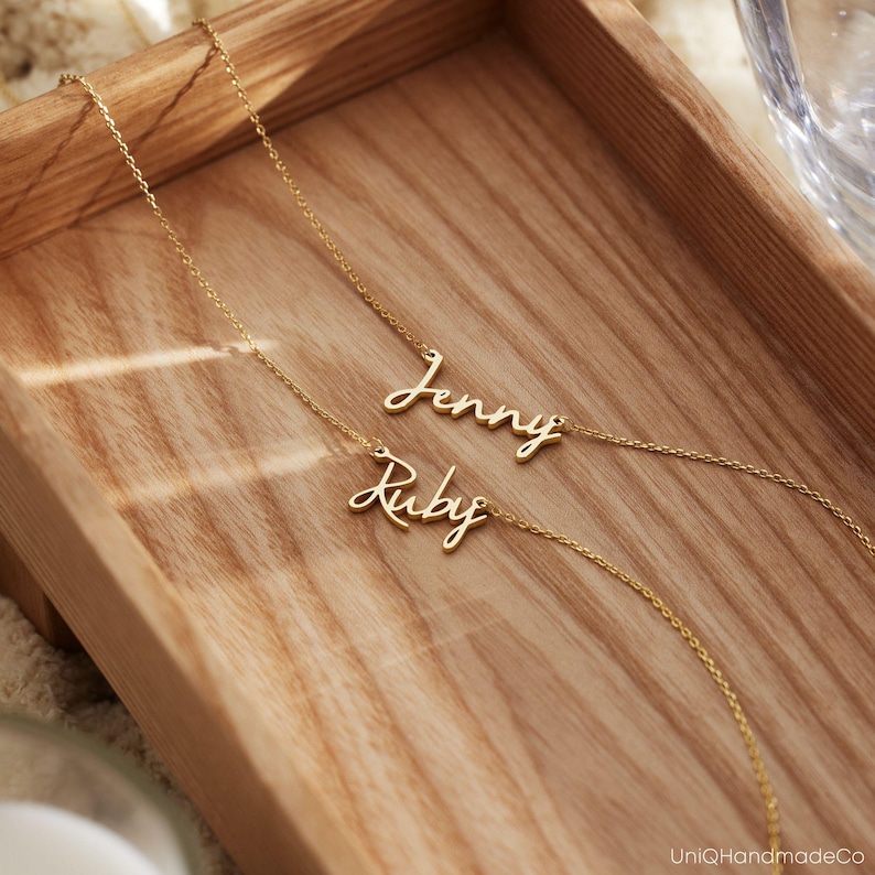 May include: Two gold necklaces with the names "Jenny" and "Ruby" written in cursive script. The necklaces are displayed on a wooden surface.