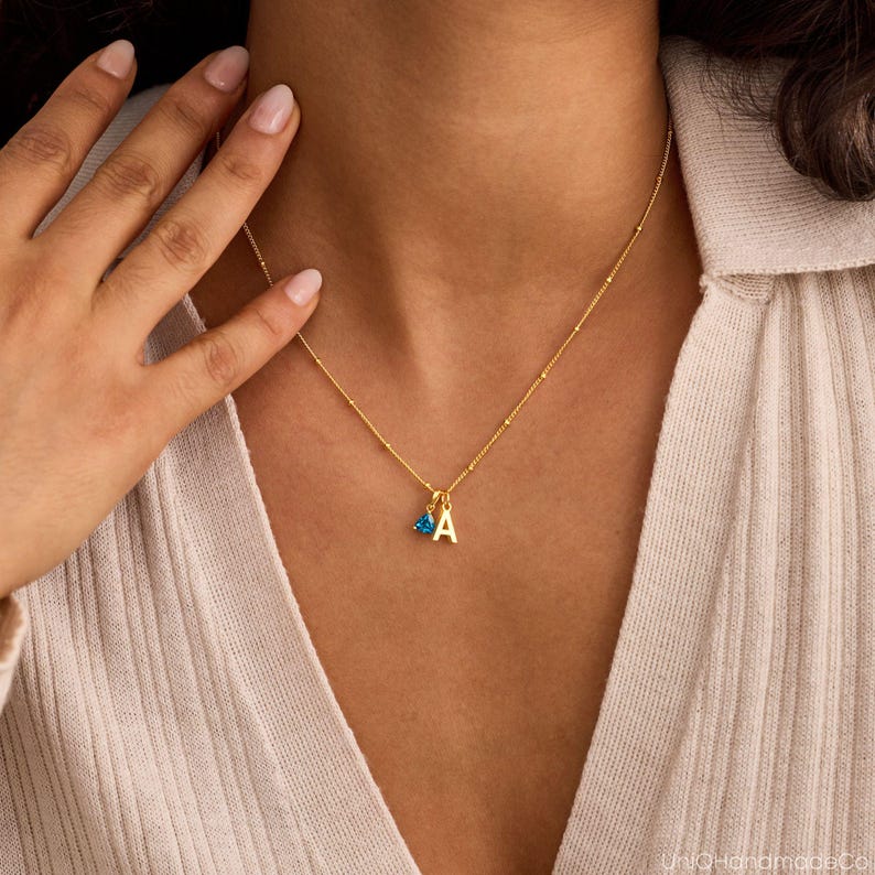 May include: Gold-colored necklace with a small pendant featuring the letter "A" and a blue gemstone. The necklace is worn around the neck, with the chain and pendant clearly visible. The background is a neutral color.