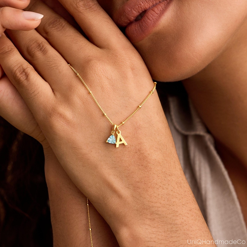 May include: A gold-colored necklace with a small pendant featuring the letter "A" and a light blue gemstone. The necklace is delicate, with small beaded details along the chain. The image is a close-up, highlighting the jewelry.