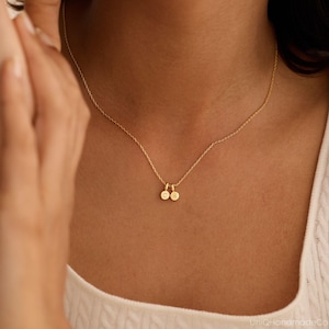 May include: A gold-toned necklace featuring a delicate chain and two small, circular pendants. Each pendant is engraved with a single letter, specifically the characters C and K, hanging side by side at the center of the chain.