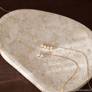 May include: Two delicate gold chains featuring small, circular charms. One chain displays four charms engraved with the letters E, I, B, and N, while the other chain features two charms engraved with the letters C and K. The chains are thin and metallic.