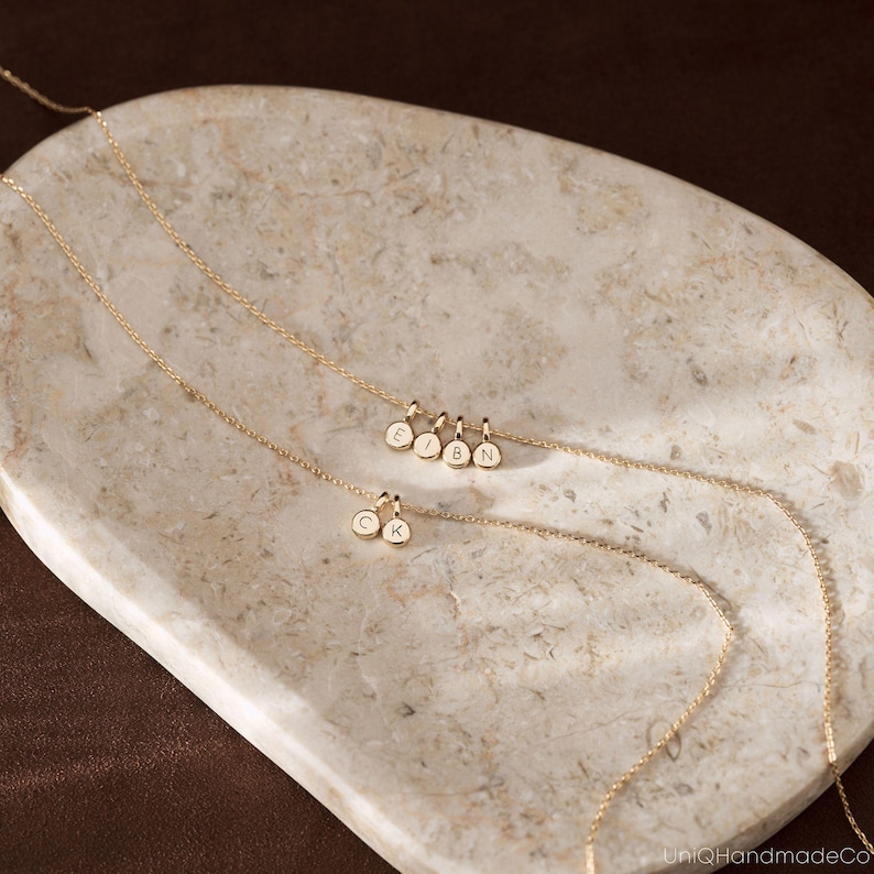 May include: Two delicate gold chains featuring small, circular charms. One chain displays four charms engraved with the letters E, I, B, and N, while the other chain features two charms engraved with the letters C and K. The chains are thin and metallic.
