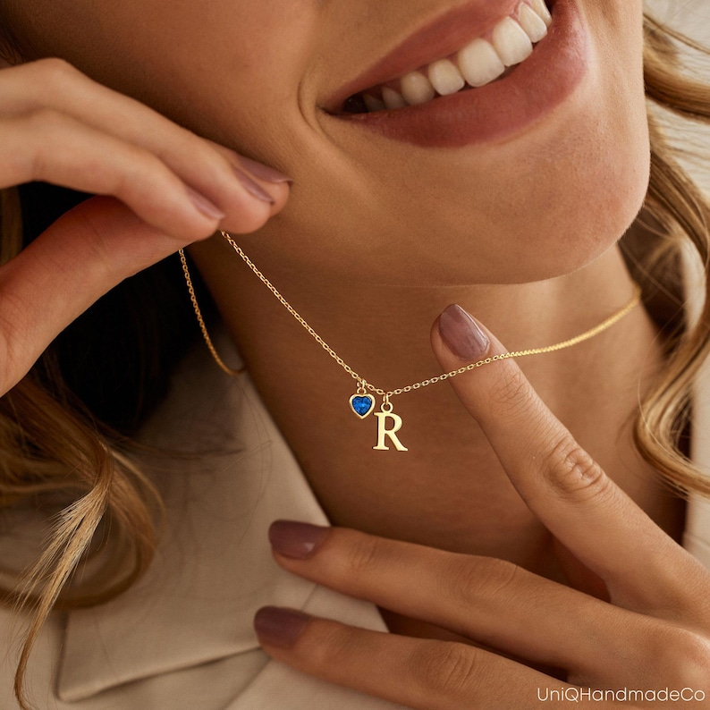 May include: A gold chain necklace with a small heart charm with a blue gemstone and a letter "R" charm.