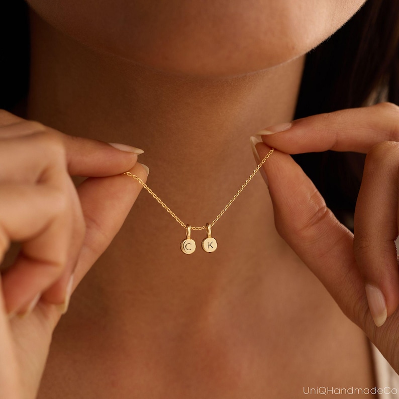 May include: A gold-toned necklace featuring a delicate chain and two small, circular charms. Each charm is engraved with a single letter, specifically the letters C and K, suspended from the chain in a side-by-side arrangement.
