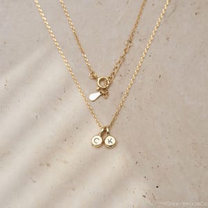 May include: A gold-toned necklace featuring a delicate chain with a spring ring clasp and an adjustable extender. Two small, circular gold-toned charms hang from the center, each engraved with a single uppercase letter, specifically C and K.