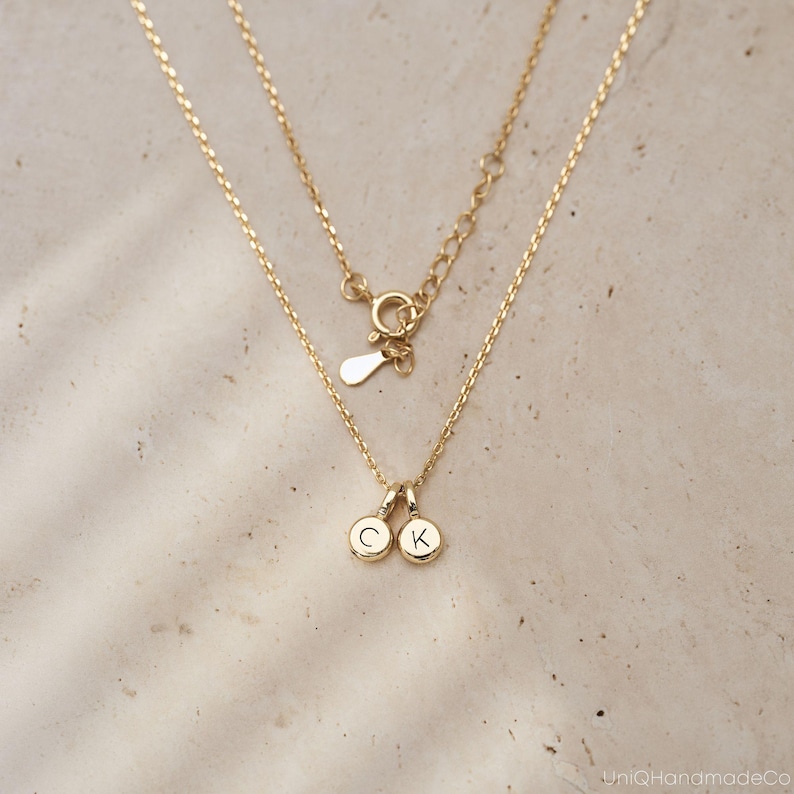 May include: A gold-toned necklace featuring a delicate chain with a spring ring clasp and an adjustable extender. Two small, circular gold-toned charms hang from the center, each engraved with a single uppercase letter, specifically C and K.