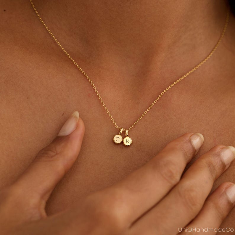May include: A delicate gold necklace with two small, round pendants. One pendant displays the letter "C", the other "K". The necklace is worn around a person's neck, with the chain and pendants in clear focus.