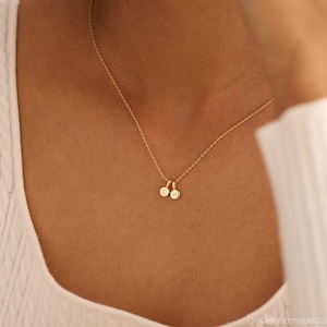 May include: A gold-toned necklace featuring a delicate chain and two small, circular pendants. Each pendant is engraved with a single letter, specifically C and K, hanging centrally from the chain. The necklace rests against a white textured fabric.