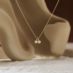 May include: A gold-toned necklace featuring a delicate chain and two small, circular charms. Each charm is engraved with a single uppercase letter, specifically C and K, suspended from the center of the chain in a simple, minimalist arrangement.
