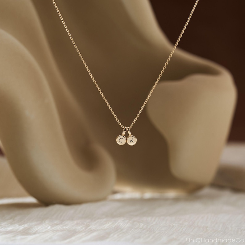 May include: A gold-toned necklace featuring a delicate chain and two small, circular charms. Each charm is engraved with a single uppercase letter, specifically C and K, suspended from the center of the chain in a simple, minimalist arrangement.