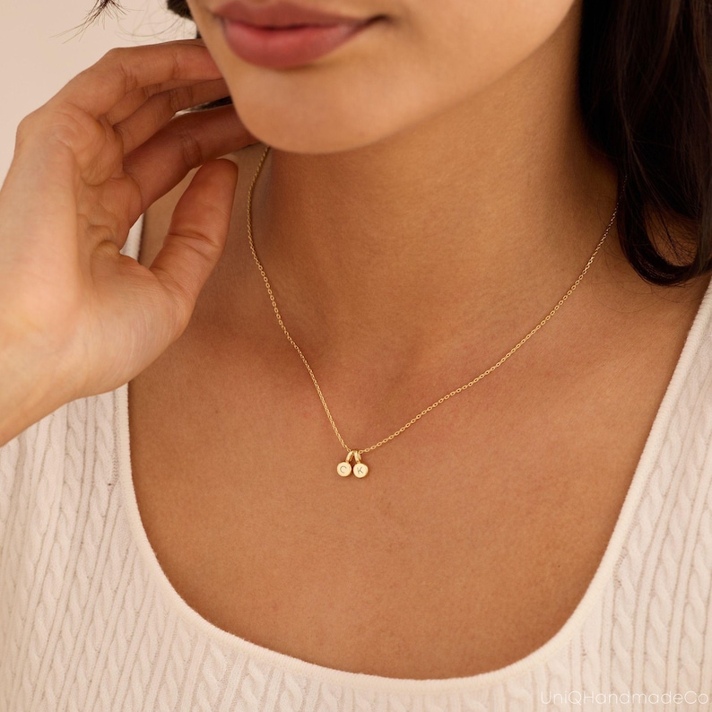 May include: A gold-toned chain necklace featuring two small, circular charms. Each charm is engraved with a single letter, specifically the characters C and K, suspended from a delicate link chain that rests against the collarbone area.