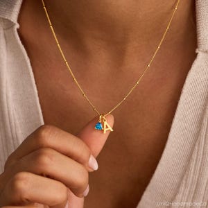 May include: A gold-colored necklace with a delicate chain and small beaded accents. The necklace features a pendant with the letter "A" and a small, triangular blue gemstone. The necklace is being held up by a person's finger.