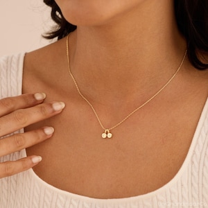 May include: A gold-toned chain necklace featuring two small, circular charms. Each charm is engraved with a single letter, specifically the letters C and K, hanging from the center of the delicate link chain against a white textured fabric.