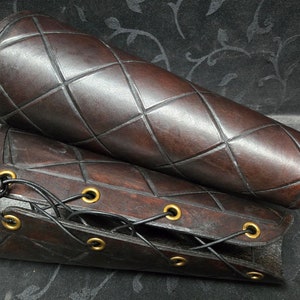 Puede incluir: Un par de protectores de brazo de cuero marrón con un diseño acolchado y ojales de latón. Los protectores están unidos con un cordón negro.