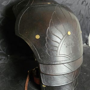 Peut inclure: Une paire de pièces d'armure d'épaule en cuir noir avec un motif d'arbre et des accents dorés. L'armure est conçue pour protéger les épaules et le haut des bras.