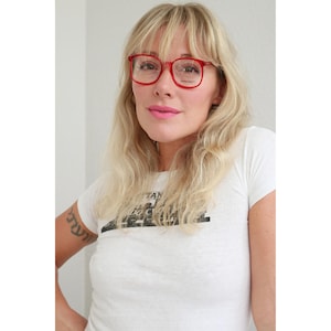 Puede incluir: Gafas rojas con montura cuadrada. Las gafas tienen un diseño audaz y clásico. La imagen muestra las gafas en una persona. La persona lleva una camiseta blanca con un estampado gráfico.