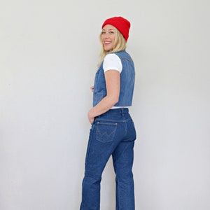 May include: A woman wearing a blue denim vest, blue jeans, and a red beanie. She is standing in front of a white wall.