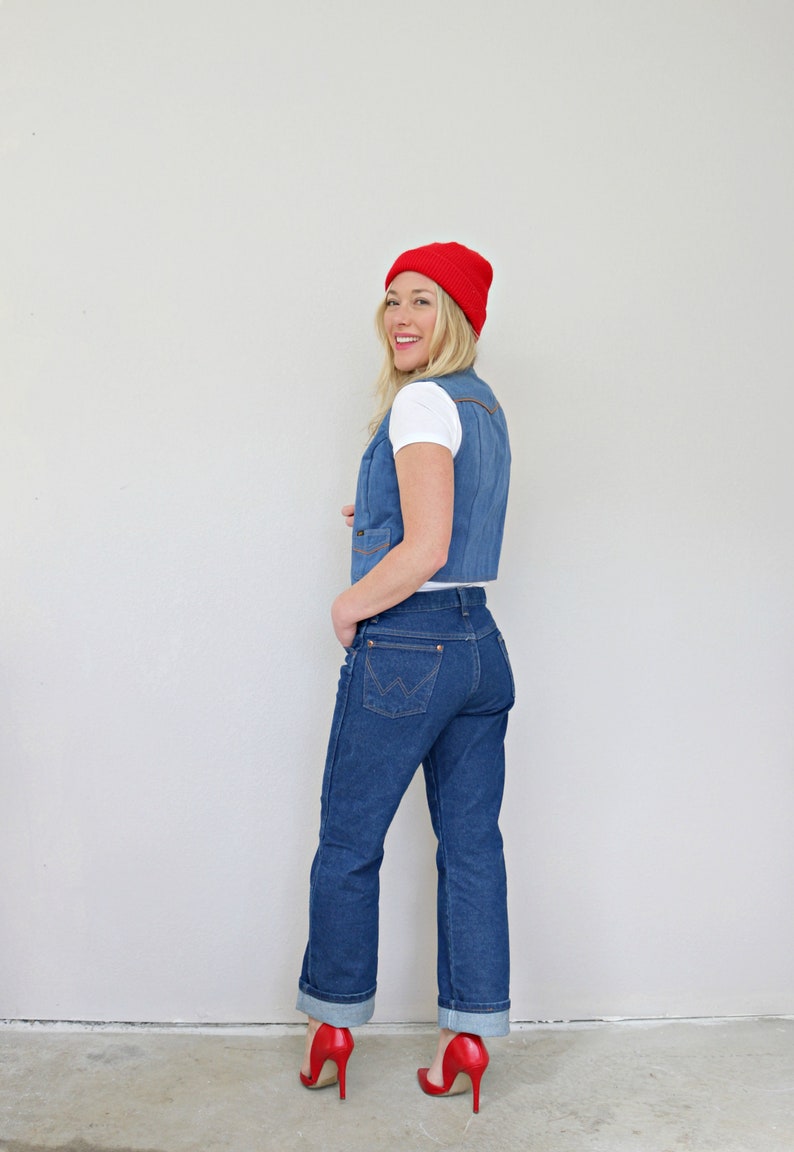 May include: A woman wearing a blue denim vest, blue jeans, and a red beanie. She is standing in front of a white wall.