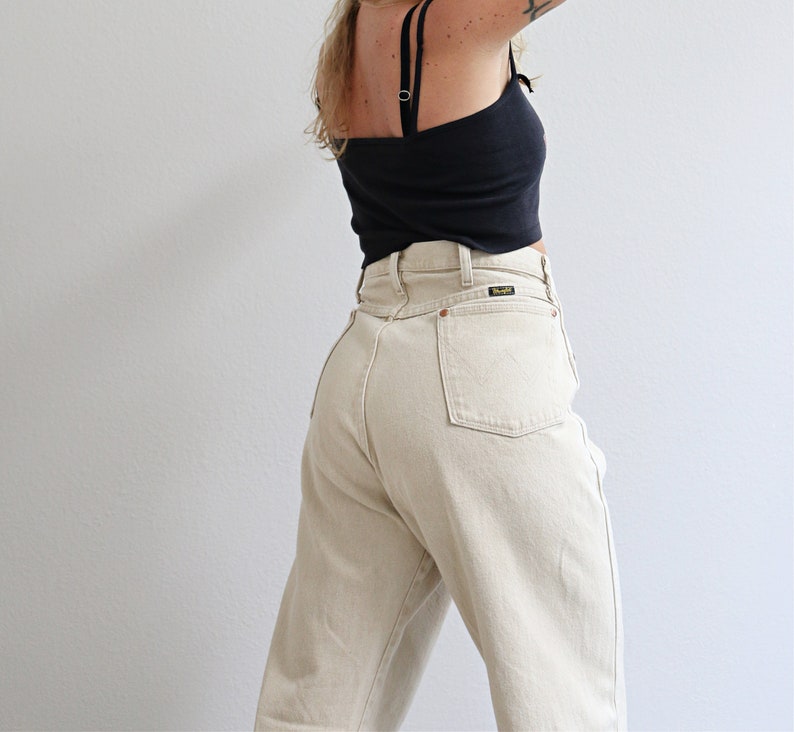 May include: A pair of high-waisted, light beige jeans with a back pocket featuring a small brown leather patch with the word "Wrangler" printed on it.