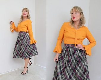 Könnte beinhalten: Ein orangefarbenes Oberteil mit Glockenärmeln und ein karierter Rock. Der Rock hat ein Muster aus schwarzen, weißen und bordeauxfarbenen Linien. Das Outfit wird mit schwarzen Absätzen kombiniert. Der Rock reicht bis zu den Knien.