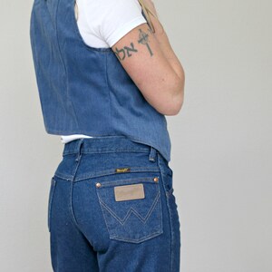 May include: A woman wearing a blue denim vest over a white t-shirt and blue denim jeans with a brown leather patch on the back pocket. The jeans have a high waist and a straight leg.