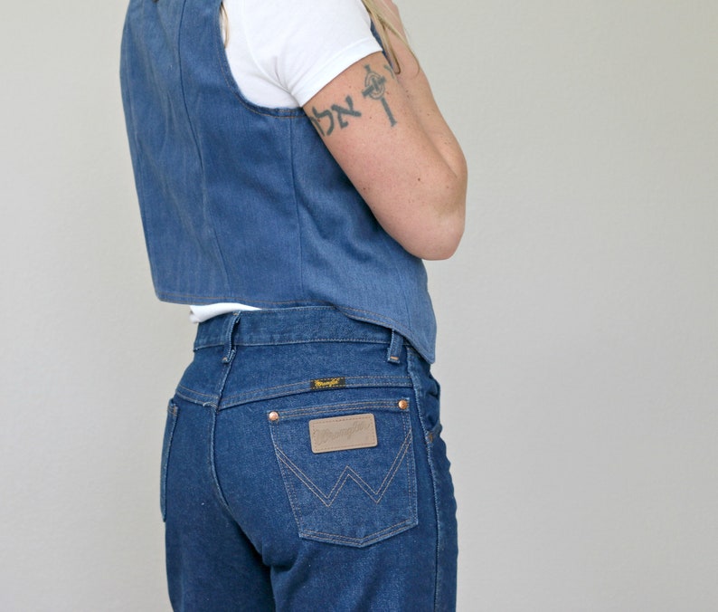 May include: A woman wearing a blue denim vest over a white t-shirt and blue denim jeans with a brown leather patch on the back pocket. The jeans have a high waist and a straight leg.