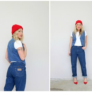 May include: A woman wearing a red beanie, a white t-shirt, a denim vest, and blue jeans. She is standing in front of a white wall.