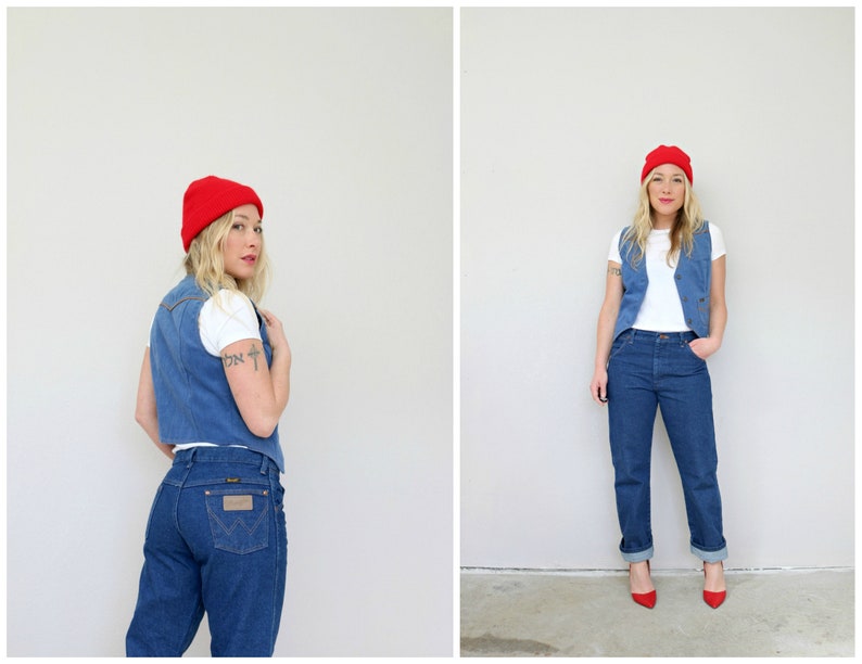 May include: A woman wearing a red beanie, a white t-shirt, a denim vest, and blue jeans. She is standing in front of a white wall.