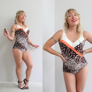 May include: A vintage one-piece swimsuit featuring a brown floral pattern with white and pink flowers. The swimsuit has a white top with orange trim and a V-neck design. The straps are white, and the swimsuit is a classic retro style.
