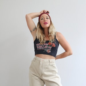 May include: A woman wearing a black tank top with a Harley Davidson logo and red roses, a burgundy beanie, and beige high-waisted jeans.