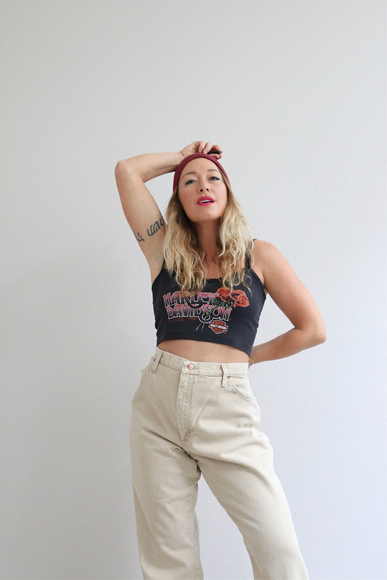 May include: A woman wearing a black tank top with a Harley Davidson logo and red roses, a burgundy beanie, and beige high-waisted jeans.