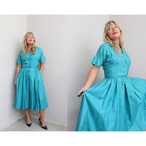 May include: A bright teal midi dress featuring a collared neckline, short sleeves with button details, and a button-down front bodice. The skirt is pleated and full, falling below the knee, with a cinched waistline and a matching fabric tie belt.