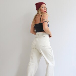 Pu&ograve; includere: Una donna indossa una canotta nera, jeans bianchi a vita alta, un berretto bordeaux e tacchi neri. &Egrave; in piedi davanti a un muro bianco.