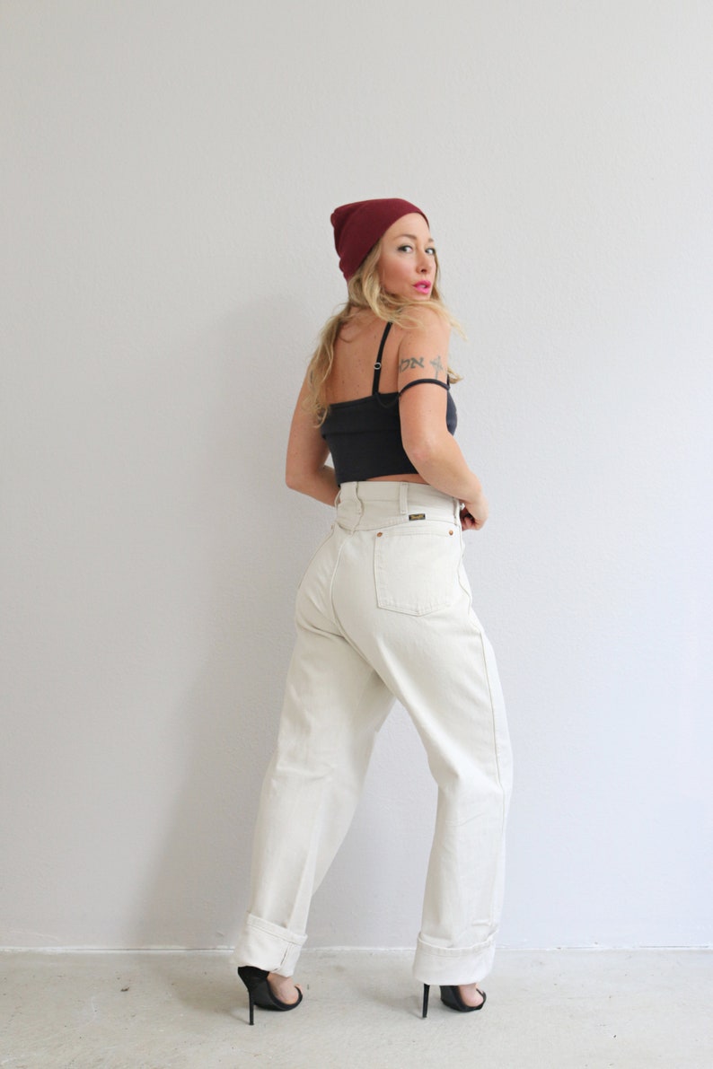 Pu&ograve; includere: Una donna indossa una canotta nera, jeans bianchi a vita alta, un berretto bordeaux e tacchi neri. &Egrave; in piedi davanti a un muro bianco.