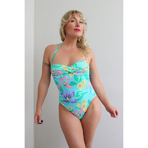 May include: A one-piece swimsuit with a halter neck and a tropical floral print. The swimsuit features a light blue background with pink, purple, yellow, and green floral designs. The swimsuit has a sweetheart neckline.