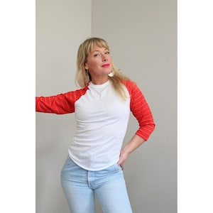 Puede incluir: Una camiseta blanca de estilo béisbol con mangas rojas. Las mangas tienen un fino patrón de rayas verticales. La camiseta se combina con vaqueros azul claro. La persona lleva pendientes y tiene el pelo rubio.