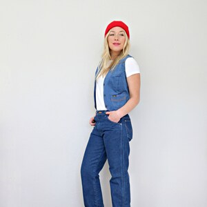 May include: A woman wearing a white t-shirt, a blue denim vest, blue jeans, red shoes, and a red beanie.