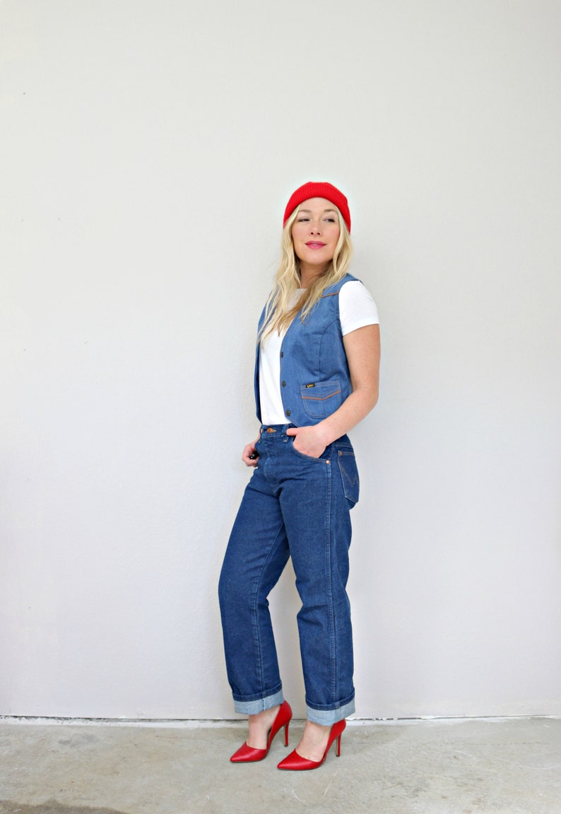 May include: A woman wearing a white t-shirt, a blue denim vest, blue jeans, red shoes, and a red beanie.