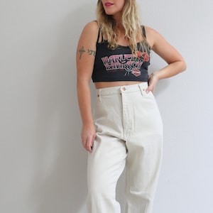 Pu&ograve; includere: Una donna indossa un top corto nero con la scritta "Harley Davidson" e un disegno floreale rosso. Indossa anche pantaloni beige chiaro a vita alta.