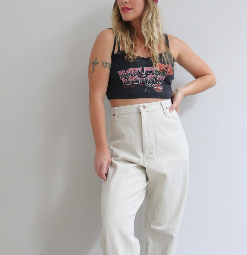 Pu&ograve; includere: Una donna indossa un top corto nero con la scritta "Harley Davidson" e un disegno floreale rosso. Indossa anche pantaloni beige chiaro a vita alta.