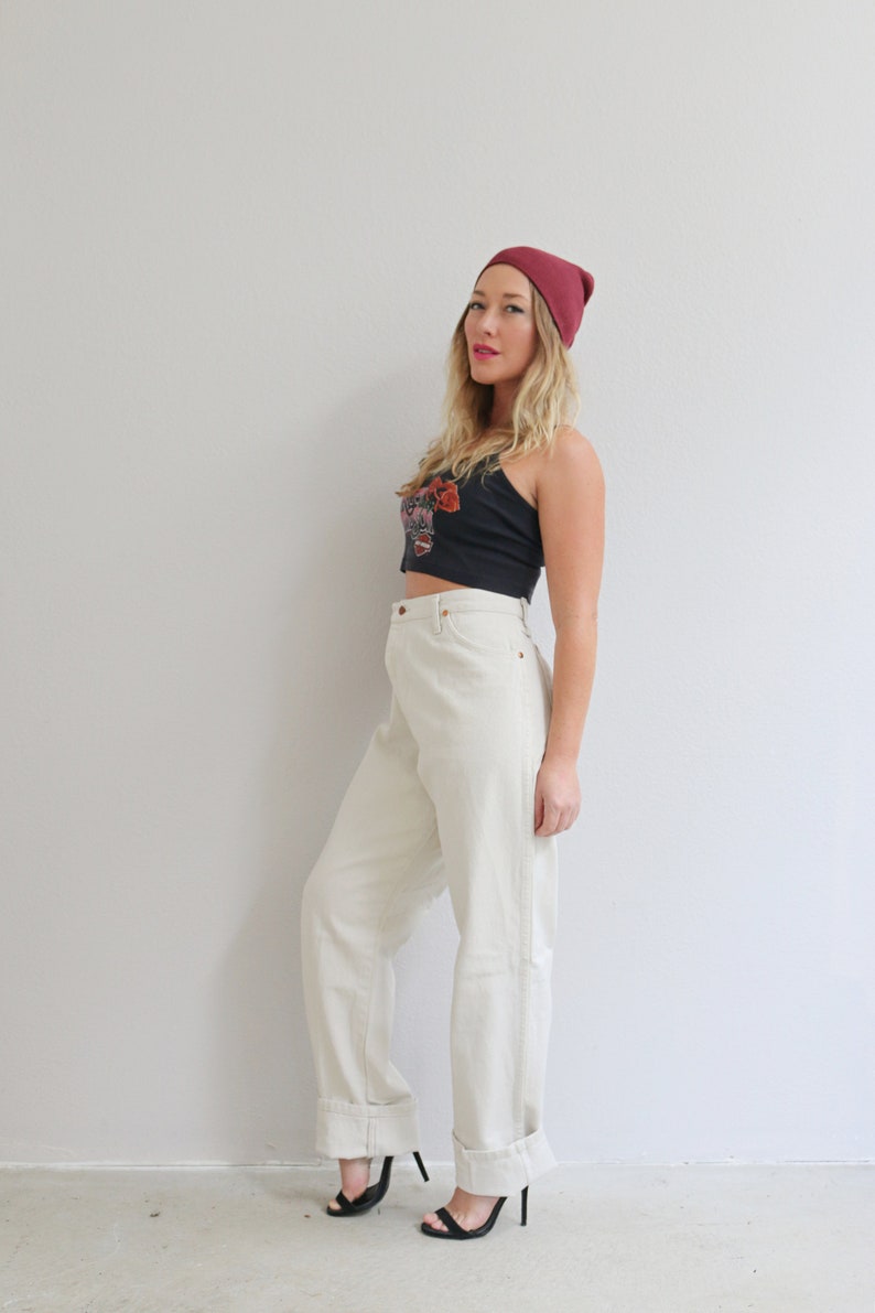 Pu&ograve; includere: Una donna indossa un paio di jeans bianchi a vita alta con orli arrotolati, un top corto nero con un motivo floreale e un berretto bordeaux.