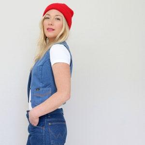 May include: A woman wearing a red beanie, a white t-shirt, a blue denim vest, and blue jeans.