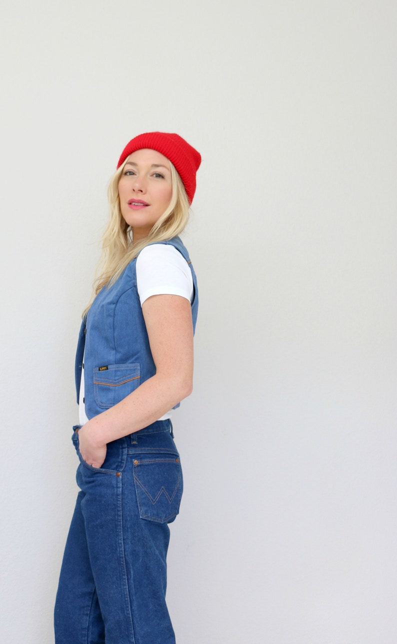 May include: A woman wearing a red beanie, a white t-shirt, a blue denim vest, and blue jeans.