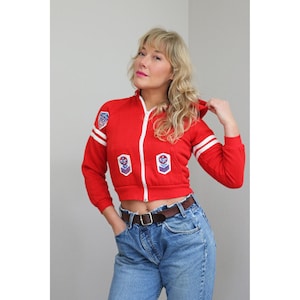May include: A red zip-up hooded jacket with white stripes on the sleeves and patches on the chest. The jacket has a white zipper and is paired with blue jeans and a brown belt. The jacket is a vintage style.