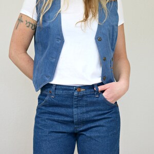 May include: A woman wearing a blue denim vest over a white t-shirt and blue jeans.