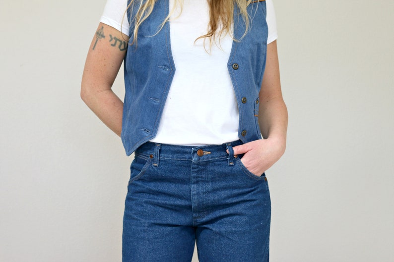 May include: A woman wearing a blue denim vest over a white t-shirt and blue jeans.