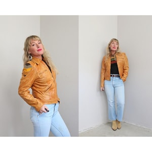 May include: A tan leather jacket with a zip-up front, multiple pockets, and shoulder patches. The jacket is paired with light blue jeans. The model is wearing a black shirt with a colorful stripe design.